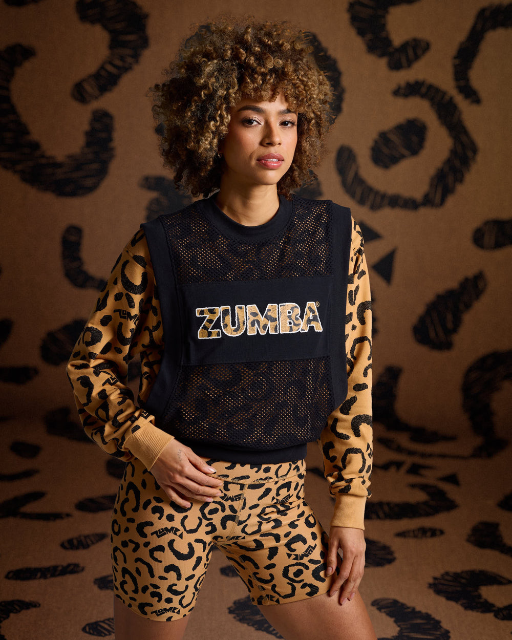 Female model wearing a black mesh Zumba tank over a cheetah print sweatshirt and matching biker shorts, against a cheetah print background.