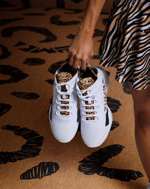 Female model holding white Zumba sneakers with cheetah print details, with a a zebra print skirt, against a cheetah print floor background.