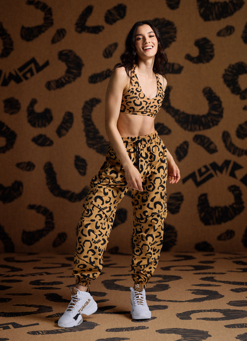 Female model wearing a matching cheetah print Zumba bra and jogger pants with white sneakers, against a cheetah print background.