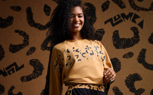 Female model wearing a tan Zumba top with mesh back and cheetah print “ZUMBA” lettering, paired with black mesh pants, against a matching cheetah print background.