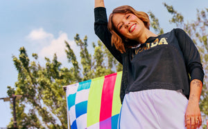 Female model wearing a black mesh top with 'Zumba' text in gold lettering, standing outdoors with a colorful flag in the background.