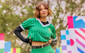 A female model in a green striped Zumba polo and matching green pants poses outdoors with colorful flags and trees in the background.