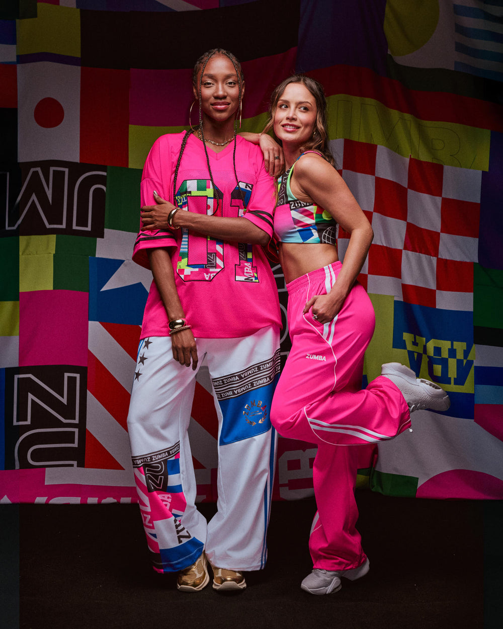 Two female models in bold pink Zumba outfits—oversized jersey, track pants, and sports bra—pose before vibrant flag backdrops.