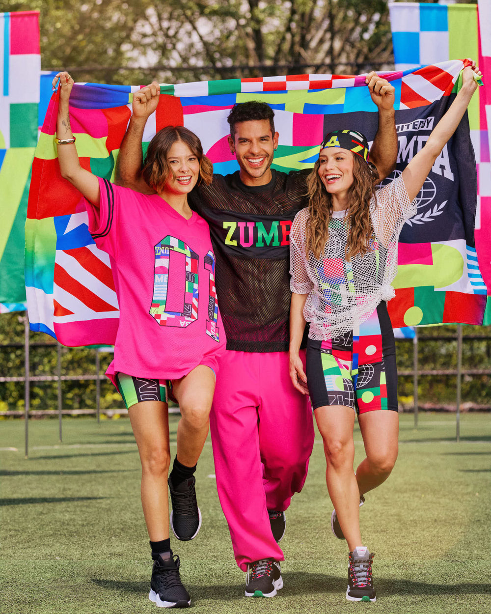 Two female models and one male model in vibrant Zumba gear celebrate on a field, holding a colorful flag with trees behind.