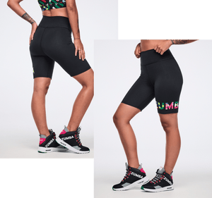 A female model in black high-waist biker shorts and Zumba sneakers poses in studio, showcasing side logo on a white backdrop.