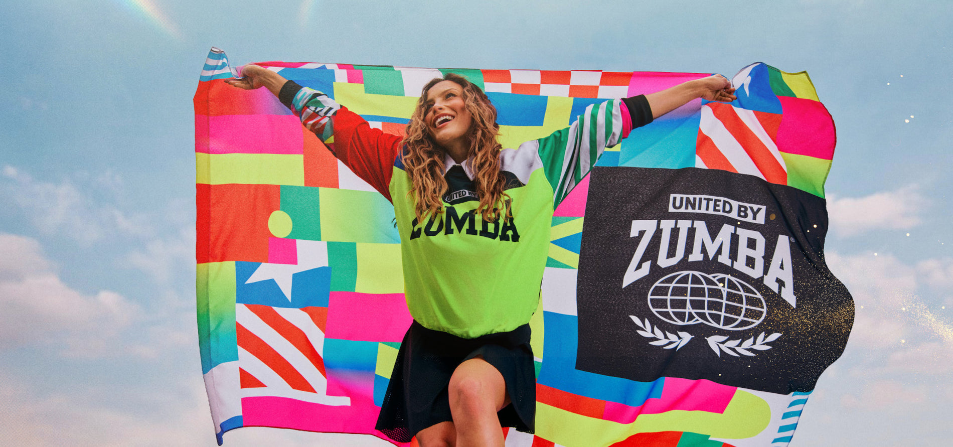 Desktop banner of a female model in a neon green Zumba jersey and black skirt raises a vibrant flag and gold specs against a bright sky backdrop.