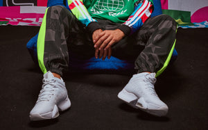 A male model in joggers with yellow stripes and white Zumba high-top sneakers sits on a blue chair against colorful backdrops.