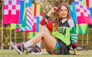 A female model in a Zumba green jersey, shorts, and black sneakers sits on grass with colorful flags in the background.