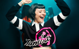 Female model smiling and dancing in a black cropped polo with a large pink Zumbito logo and black‑and‑white cursive “Zumba,” with her arms raised in a lively studio scene.