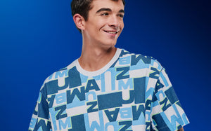 Close-up of a male model wearing a blue and white all‑over ZUMBA logo graphic top against a blue background.