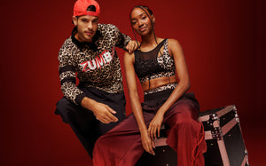 Banner collection image of a male and female model wearing coordinated Zumba activewear: The male is wearing a leopard-print Zumba top with black pants and red cap. The female is wearing a leopard-print crop top with red wide-leg pants and black sneakers, posing on a black equipment case against a red background.