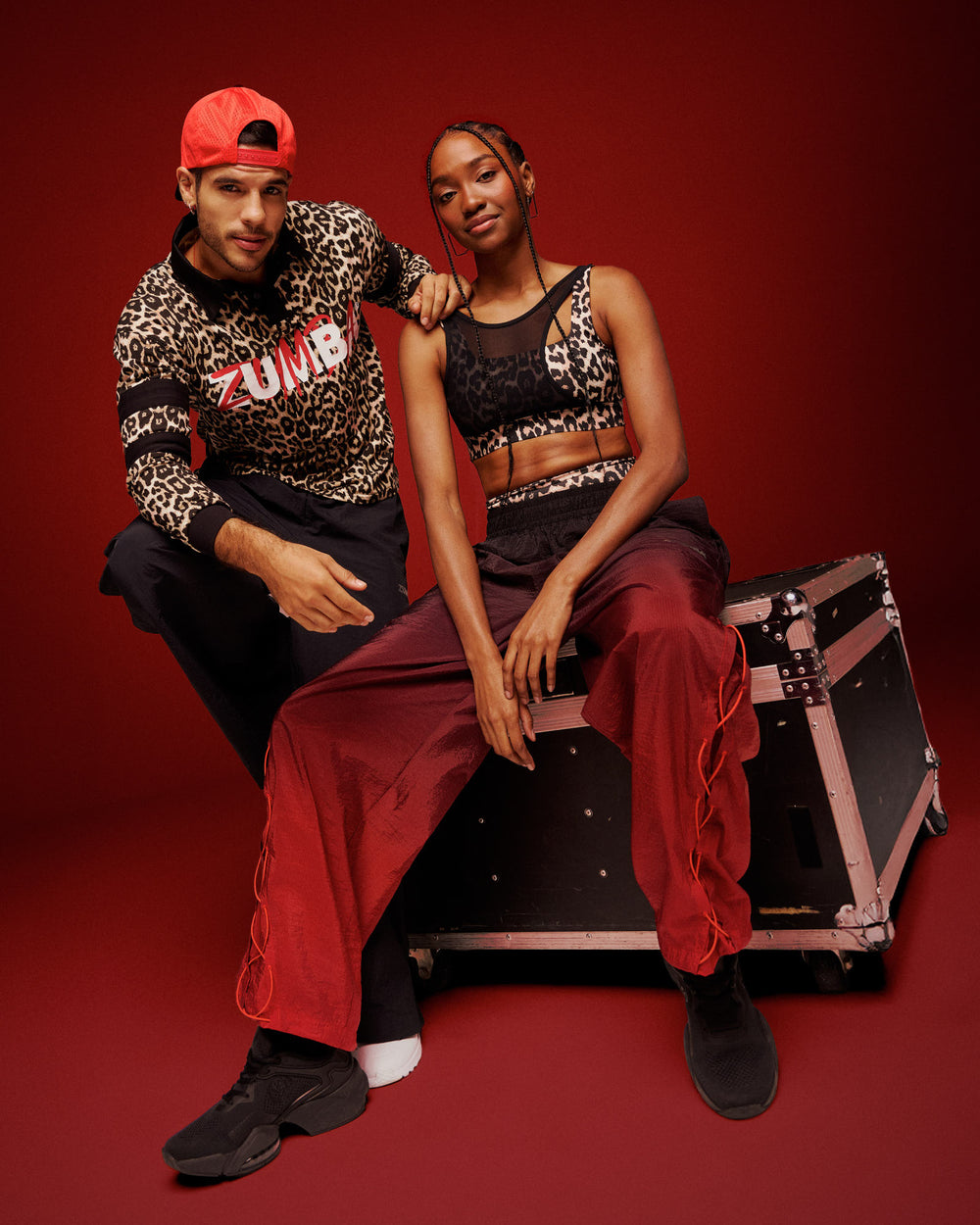 Male and female models wearing coordinated Zumba activewear: The male is wearing a leopard-print Zumba top with black pants and red cap. The female is wearing a leopard-print crop top with red wide-leg pants and black sneakers, posing on a black equipment case against a red background.