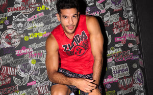 A male model wears a red ZUMBA graphic tank top against a bold black-and-white rock-inspired ZUMBA backdrop.