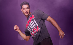 A male model poses in a dark ZUMBA graphic tee against a purple gradient background.