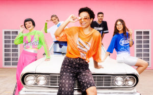 Group of five models in bright Zumba apparel gather on a white car before a pink wall, smiling and striking energetic poses.