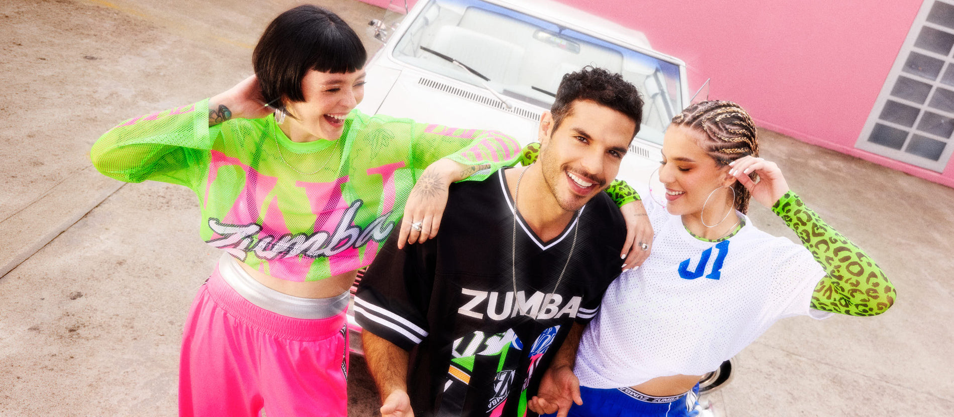Three friends pose in colorful Zumba outfits beside a white vintage car, smiling and leaning in under a pink-wall backdrop.