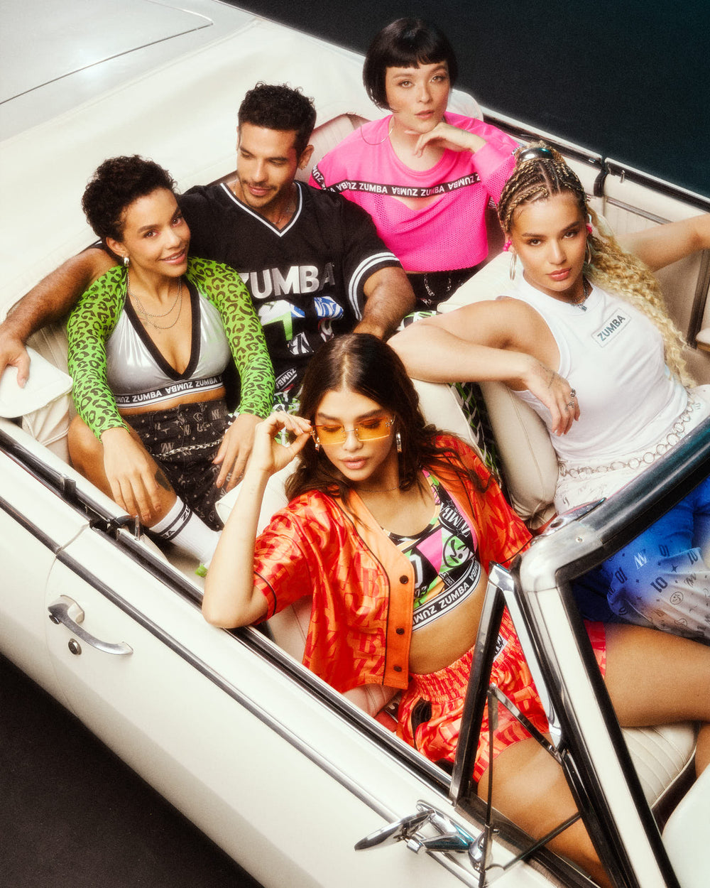 Five models sitting in a white convertible, showcasing vibrant Zumba outfits including an orange set, black jersey, pink mesh crop, silver bra with a green and black leopard print top worn as sleeves, and a white tank.