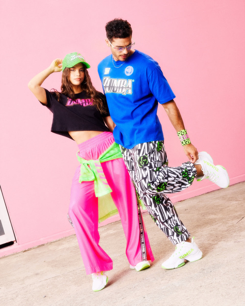 Duo posing against a pink wall: she wears a black crop tee, pink wide‑leg pants, and a green cap with a neon green top tied at the waist; he wears a blue Zumba tee, black‑white logo pants with green accents, and white‑green sneakers.