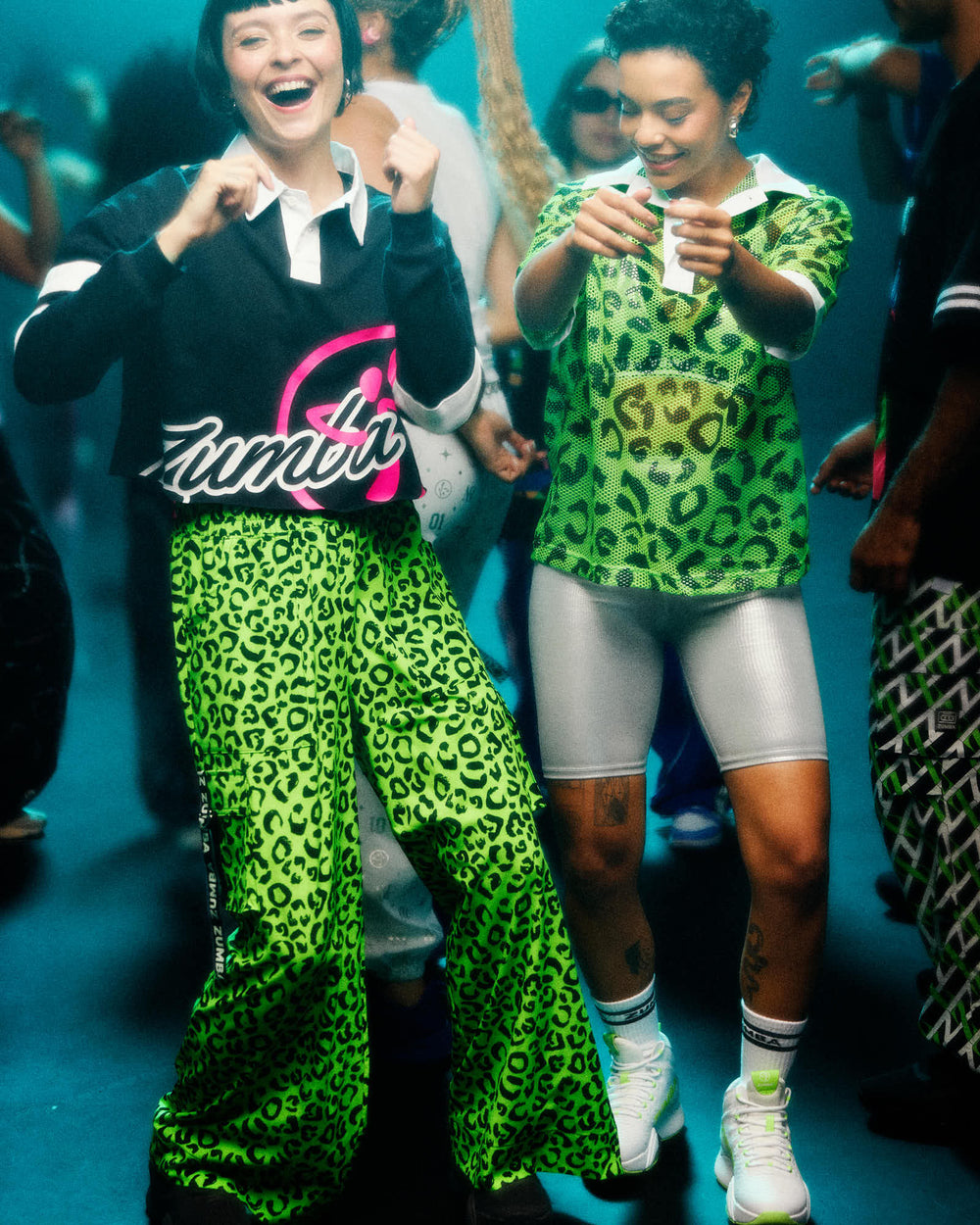 Two dancers in a teal‑lit club: left wears a black cropped polo with a pink Zumba logo and neon green leopard wide‑leg pants; right wears a green leopard mesh jersey over a silver bra with silver biker shorts and white‑green sneakers.