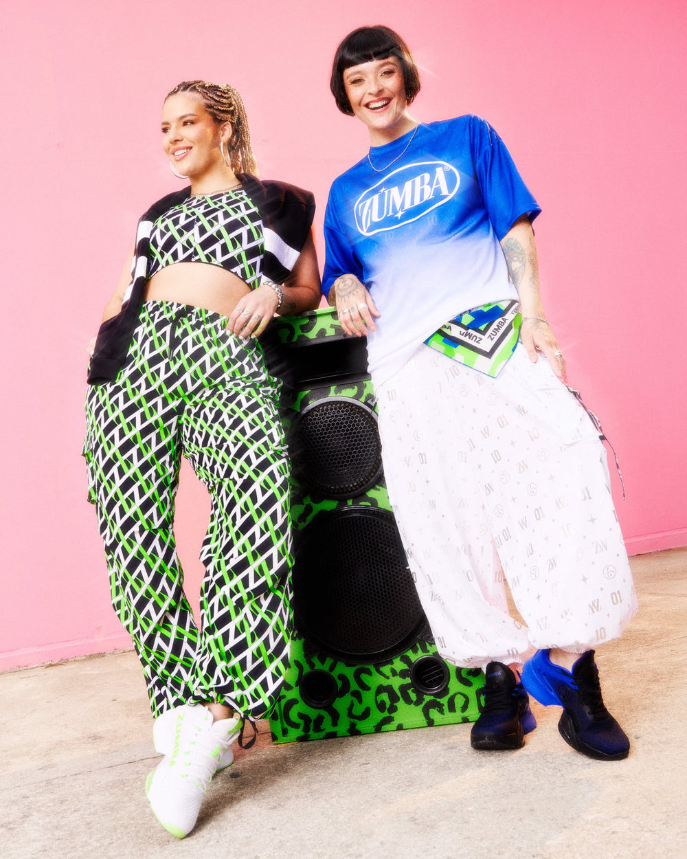 Two models standing by a green leopard speaker against a pink wall: left wears a black‑white‑green geometric set with white sneakers; right wears a blue ombré Zumba tee over white printed joggers with blue sneakers.