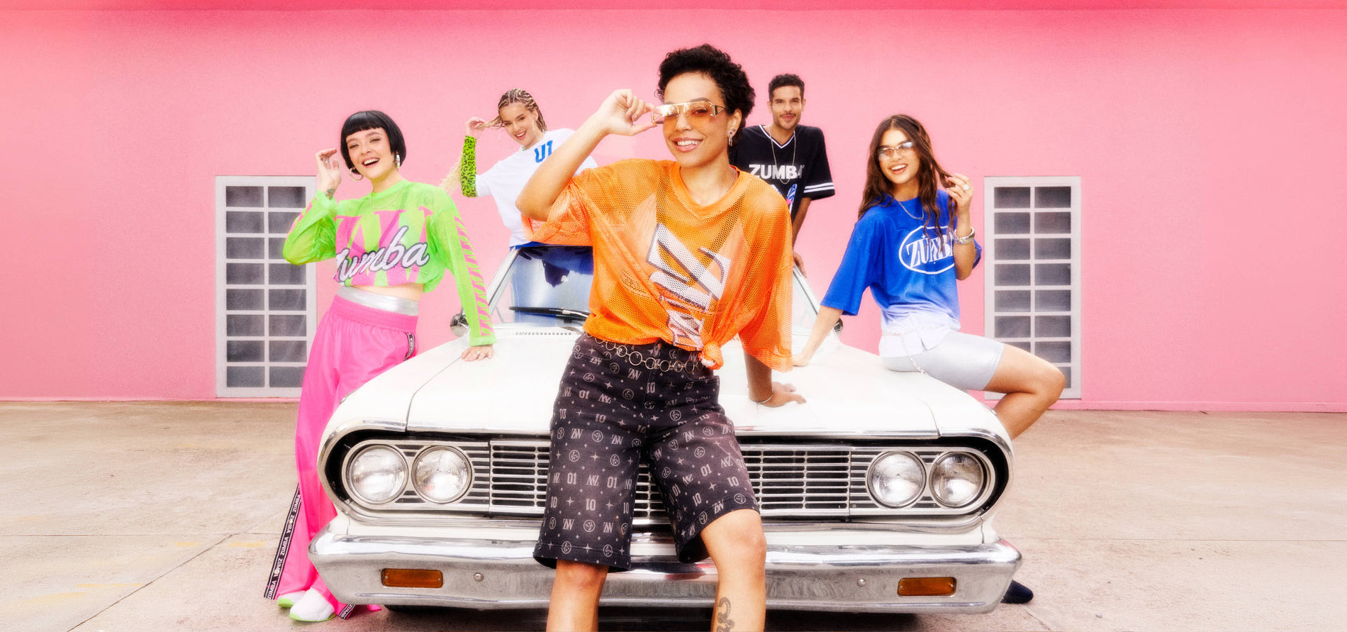 Five models pose around a vintage white car against a pink wall, showcasing colorful Zumba outfits including neon mesh tops, graphic jerseys, and silver biker shorts and black and silver patterned jorts.