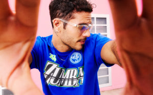 Male model with his hands on the camera and wearing a bright blue Zumba T‑shirt with bold logo graphics, photographed against a pink building.