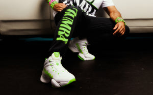 Close-up of a male model crouching by a car, showcasing white Zumba sneakers with neon green accents and black joggers with a bold green “ZUMBA” logo.