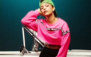 Female model leaning on a car door in a hot‑pink mesh cropped Zumba top and a green leopard cap, set against a deep teal backdrop.