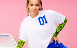 Female model in a white mesh top with a blue “01,” layered over green leopard top and paired with blue bottoms, leaning on a vintage white car in front of a pink wall.