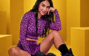 Close-up of a female model wearing a purple and black leopard print sheer top and matching biker shorts, paired with black socks and shoes against a yellow geometric backdrop.