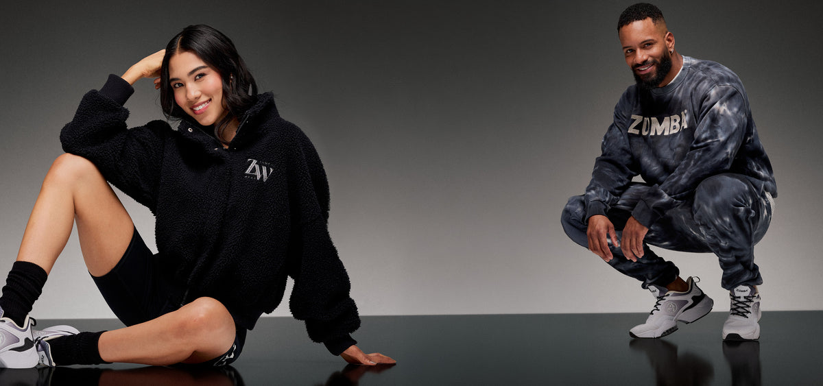 Desktop banner with a female model in a black sherpa fleece hoodie and a male model in a black and grey tie-dye 'ZUMBA' crewneck sweatshirt, both crouched against a grey gradient background.