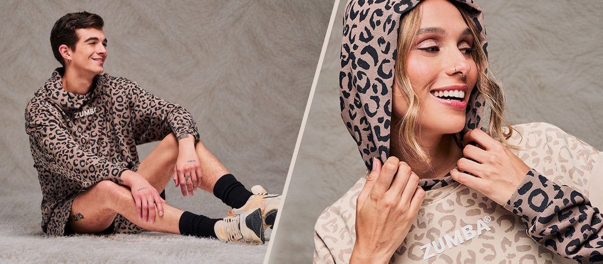 Two-panel banner split by a diagonal cream line over a taupe faux fur backdrop. On the left, a male model sits on a shaggy cream rug in a brown leopard print hooded pullover sweatshirt, matching leopard print shorts and beige shoes. On the right, a close-up of a smiling female model pulling a brown leopard print hoodie, worn under a beige sweatshirt.