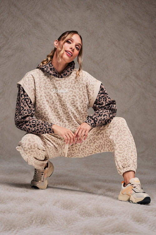 Female model crouching on a shaggy cream rug, wearing a beige leopard print sleeveless sweatshirt, layered over a brown leopard print hoodie, matching beige leopard print slouch cargo pants and beige shoes, set against a taupe faux fur backdrop.