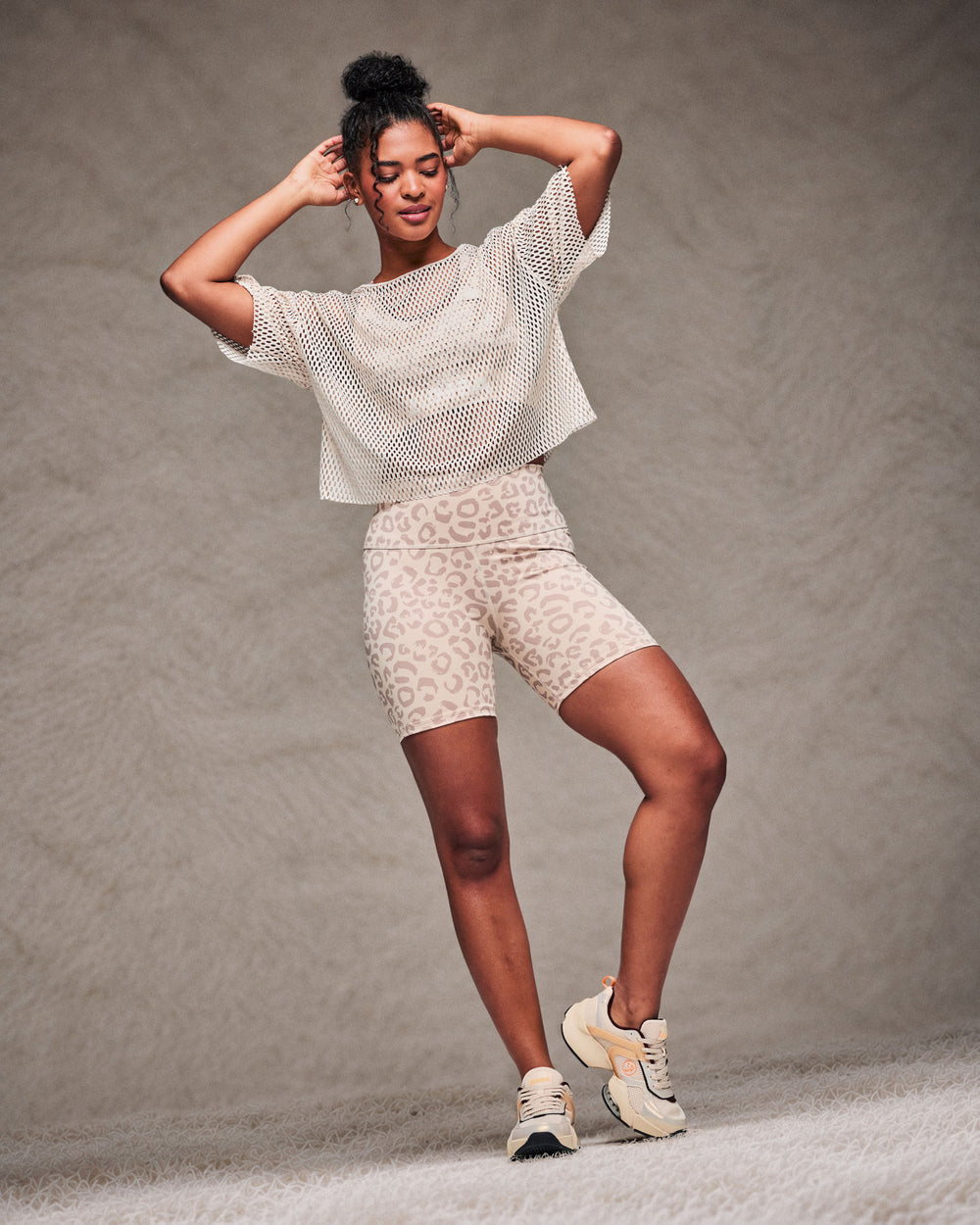 Female model mid-dance with both hands at her head, wearing a beige open-hole mesh crop top, beige leopard print high-waisted biker shorts and beige shoes on a shaggy rug and against a taupe faux fur backdrop