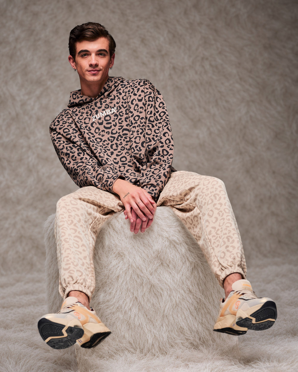 Male model perched on a shaggy white fur pouf, feet lifted, wearing a brown leopard print hooded pullover sweatshirt, beige leopard print slouch cargo pants and beige shoes against a taupe faux fur backdrop.