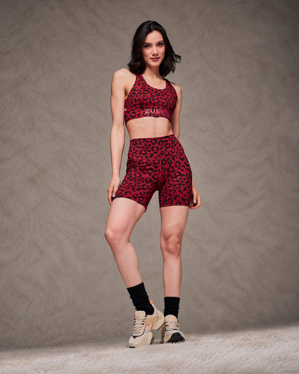 Full view of a female model in a dark red leopard print racerback bra with red 'ZUMBA' text on the underband and matching high-waisted biker shorts, paired with black socks and beige shoes. Taupe faux fur backdrop.