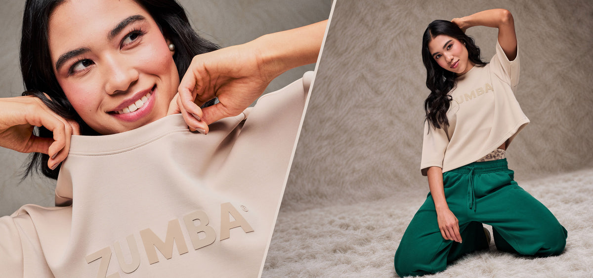 Two-panel banner split by a thin diagonal cream line. On the left, a close-up of a smiling female model tugging up the crew neckline of a beige drop shoulder crop top with large raised tonal 'ZUMBA' text. On the right, the same model kneels on a shaggy cream rug in the beige crop top and teal green pants, against a taupe faux fur backdrop.