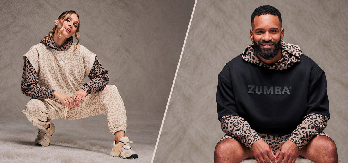 Two-panel banner divided by a diagonal cream line, both sides set against a taupe faux fur backdrop. On the left, a female model crouches in a beige leopard print sleeveless sweatshirt, layered over a brown leopard print hoodie, with matching beige leopard print slouch cargo pants and beige shoes. On the right, a seated male model wears a black oversized top with dark 'ZUMBA' text over a brown leopard print hoodie and matching shorts.