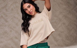 Female model with one arm raised behind her head, wearing a beige drop crop top with tonal 'ZUMBA' text over teal green pants with a drawcord waist. Taupe faux fur backdrop.