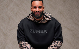 Smiling male model in a black oversized drop shoulder top with dark 'ZUMBA' text, layered over a brown leopard print hoodie with the hood down and printed sleeves showing below. Taupe faux fur backdrop.