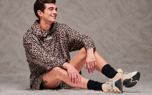 Male model seated on a shaggy cream rug, knees up, wearing a brown leopard print hooded pullover sweatshirt, matching shorts, plus black socks and beige platform shoes. Taupe faux fur backdrop.