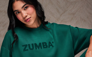 Close-up of a female model, head tilted, wearing a teal green crop top with large raised tonal 'ZUMBA' text across the chest. Taupe faux fur backdrop.