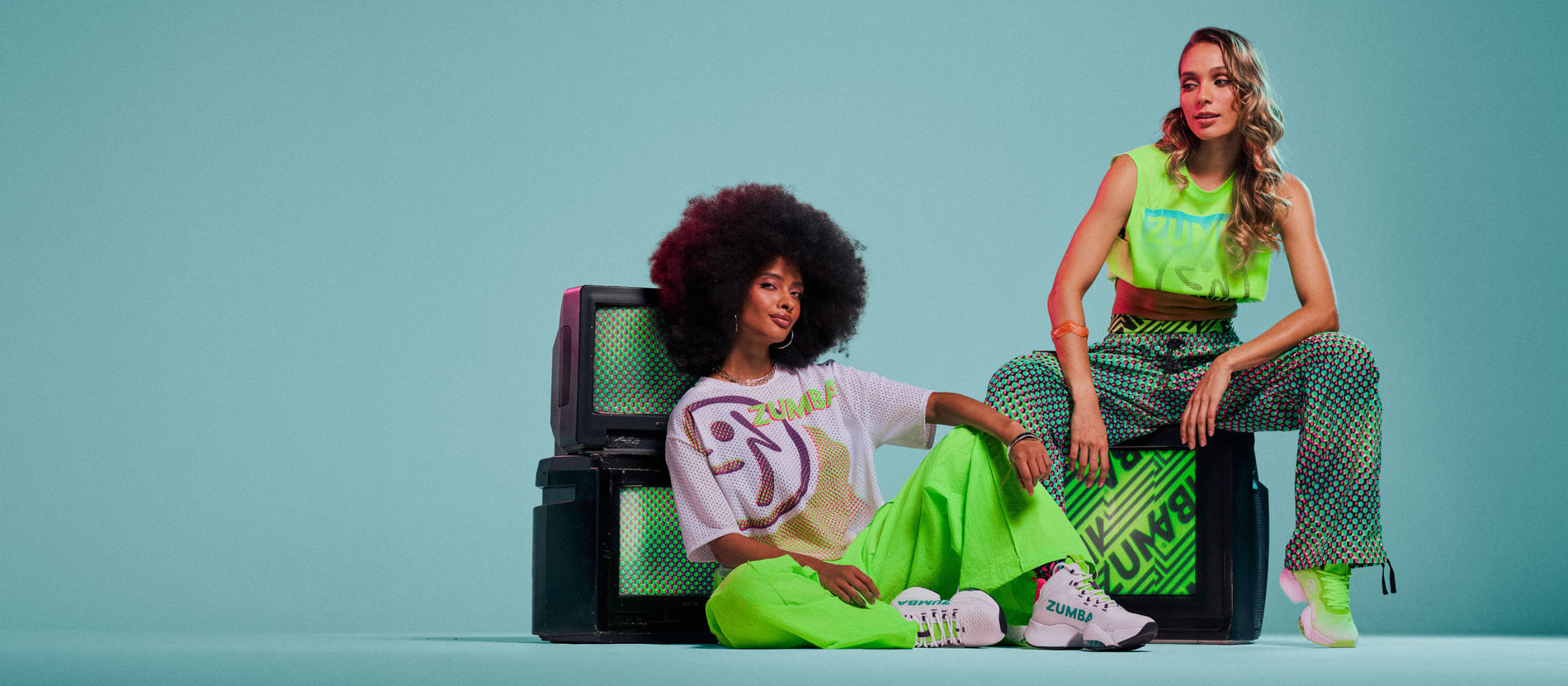 Two female models seated on vintage TVs on an aqua background, wearing a white Zumbito graphic tee with green pants, and a green tank with dot-print green pants.