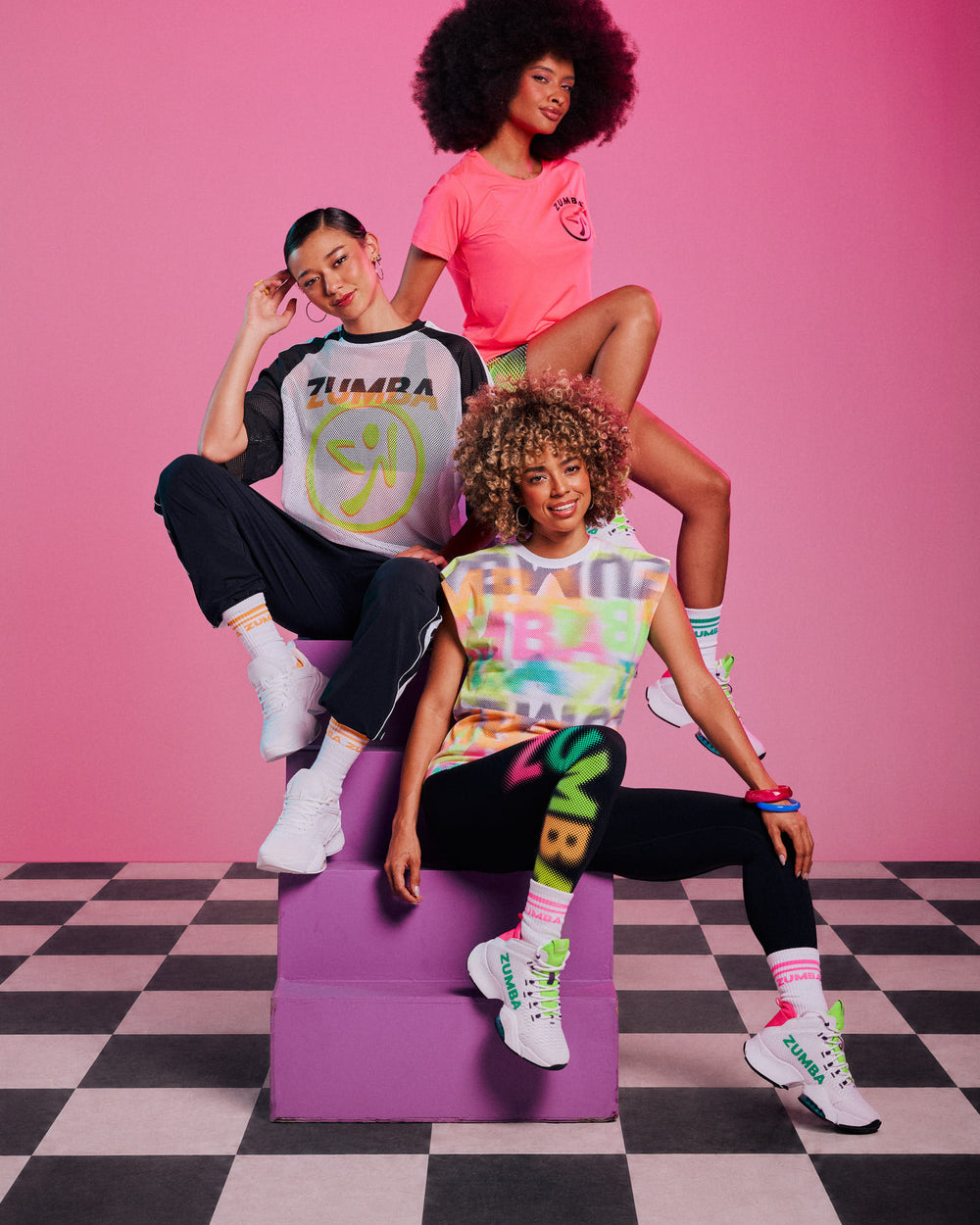 Group of three female models posed on purple stairs against a pink and checkerboard floor background, wearing a mesh Zumbito top, a pink tee, multi-color  extended shoulder tank paired with biker shorts, track pants and leggings.