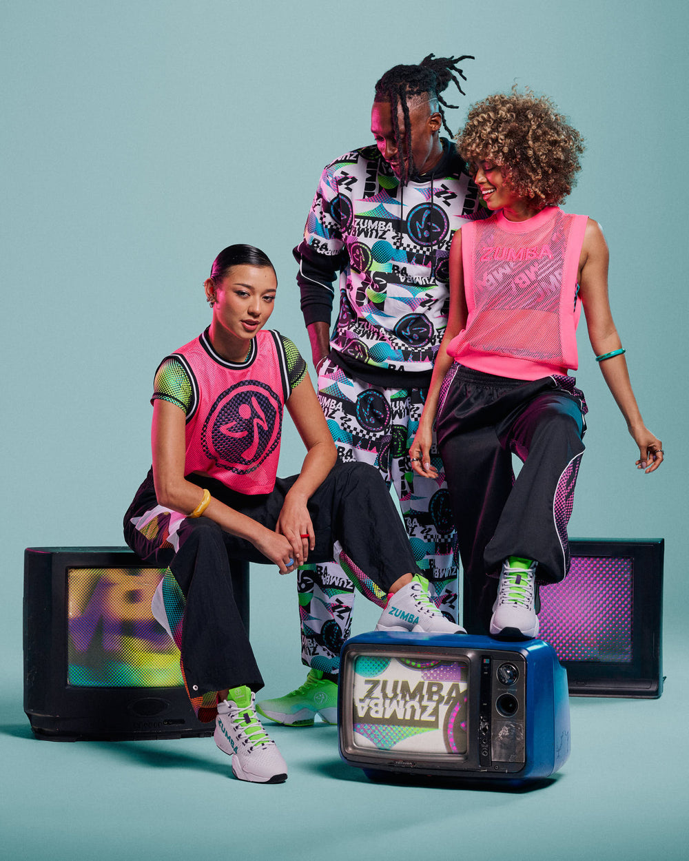 Group of three male and female models gathered around vintage TVs against an aqua background, wearing a pink mesh jersey, a black and white checkered sweatsuit (male), and a pink mesh tank with black pants.