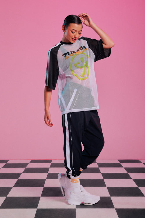 Female model wearing a white mesh top with a Zumbito graphic, paired with black joggers featuring a white side stripe, white and orange 
