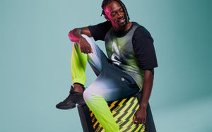 Male model seated on a lightning bolt-patterned television against an aqua background, wearing a black and green dot-pattern mesh top and grey-to-green ombre joggers.