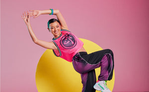 Female model in a dynamic pose against a pink background with a yellow circle, wearing a pink mesh jersey with a Zumbito graphic and black pants with a pink dot-gradient panel.