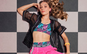 Female model lying on a black and white checkerboard floor, wearing a black mesh button-up top over a pink, teal, and black striped 