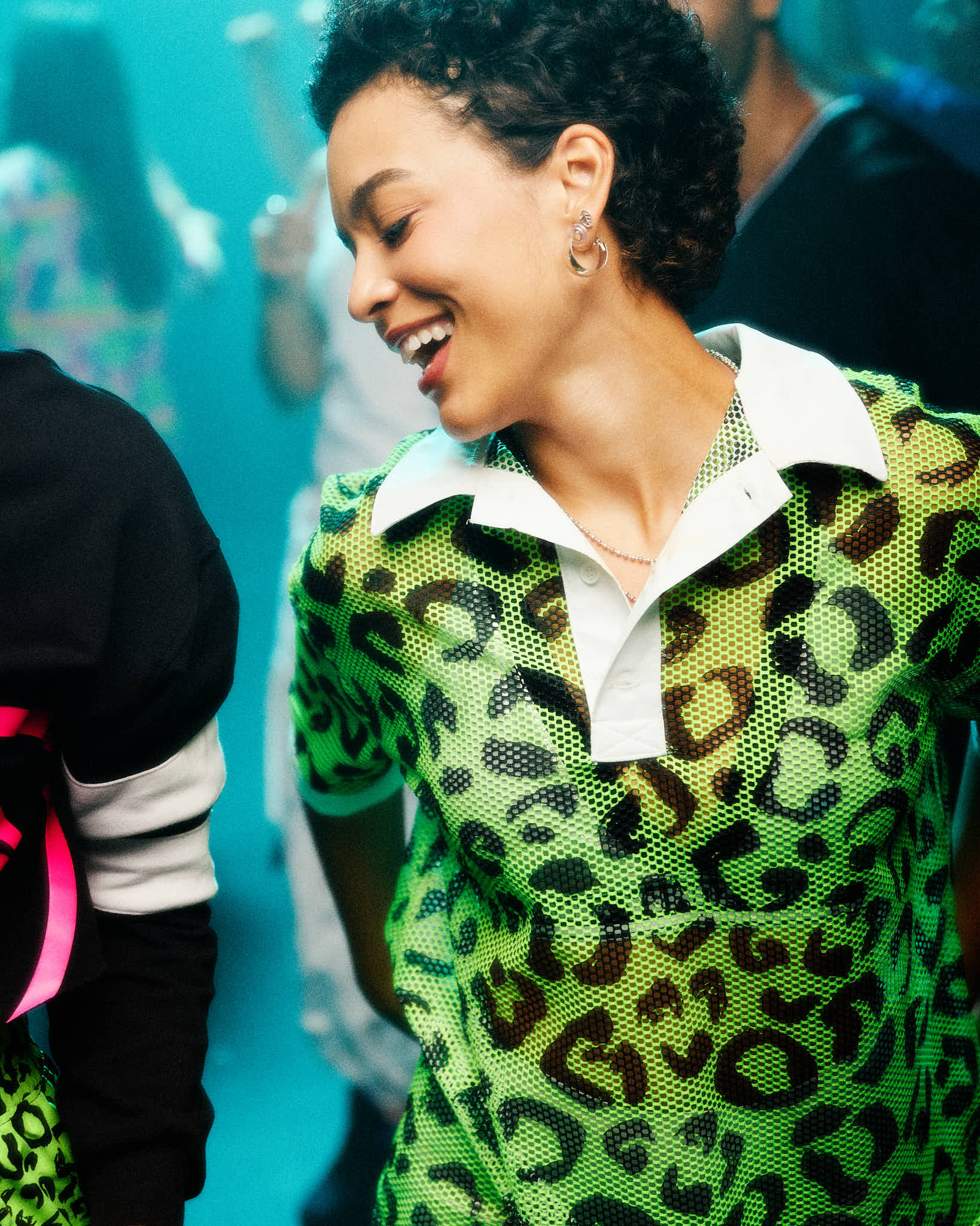 A female model wearing a green leopard-print mesh polo with a white collar, smiling in a crowded indoor setting with a blue-toned background.